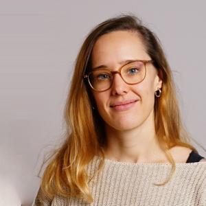 Junge Person mit langen Haaren und Brille, die lächelnd in die Kamera blickt.